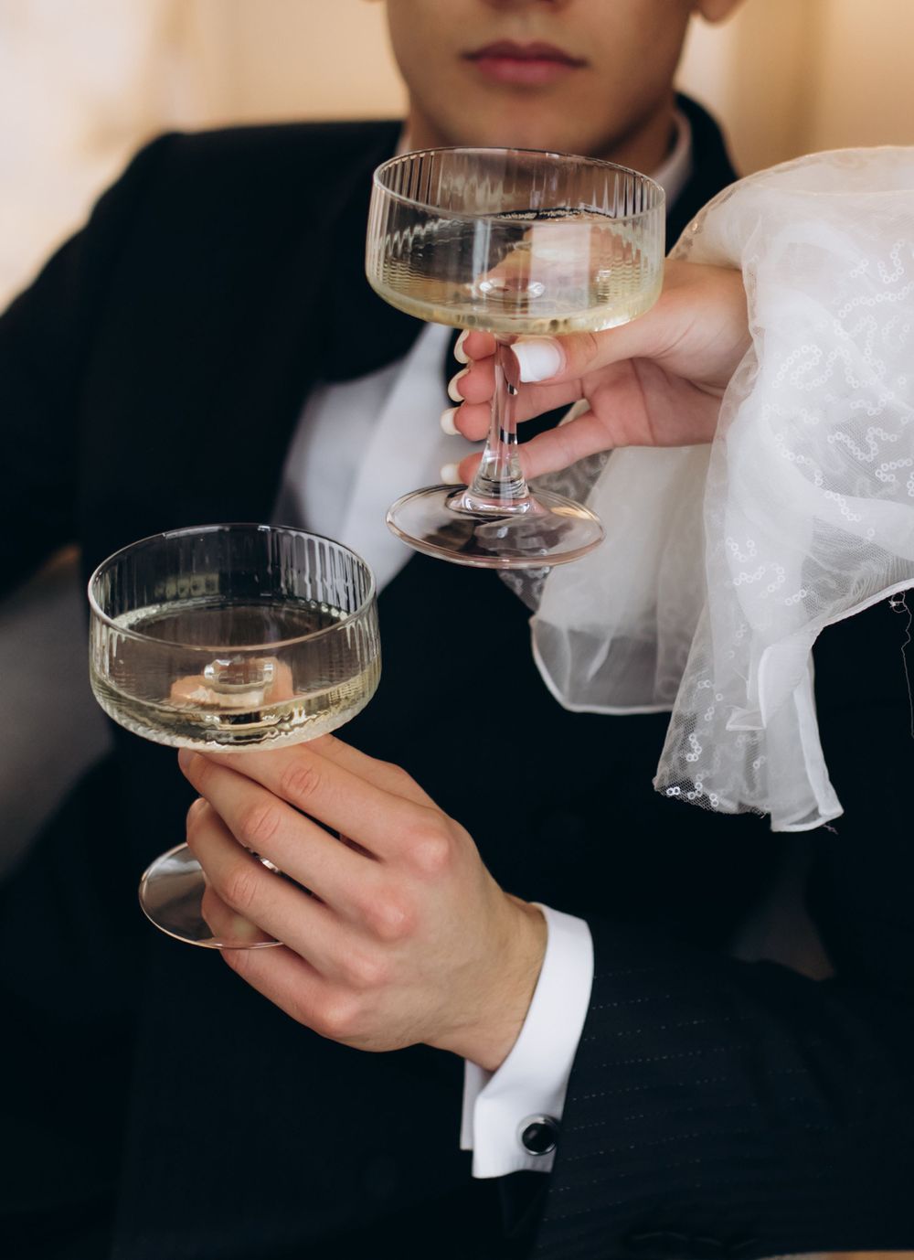 Hands holding champagne coupe glasses with white lace sleeve and formal attire during an elegant celebration.