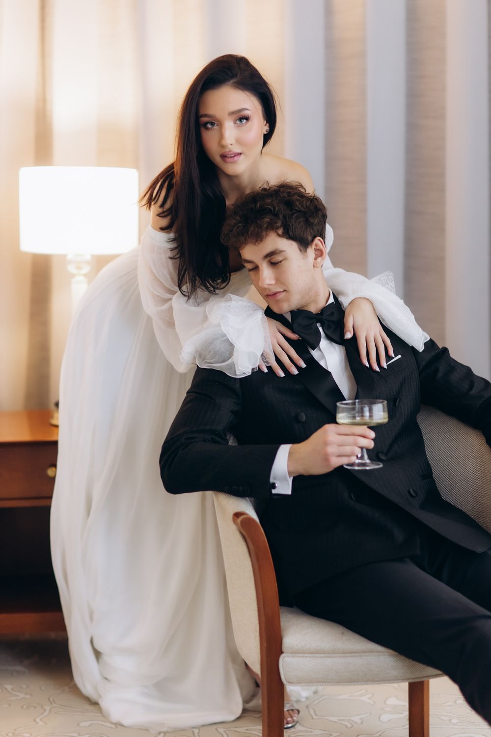 Elegant couple in wedding attire pose together, with the figure in black suit holding champagne while seated in chair.