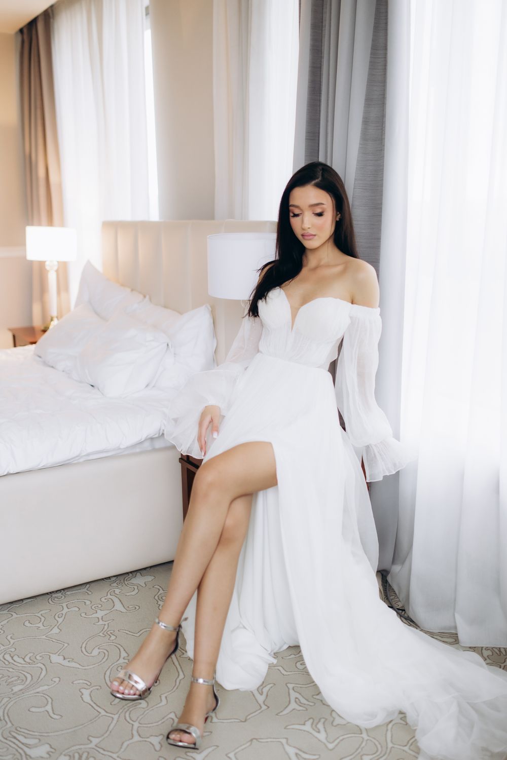 An elegant white off-shoulder wedding dress with flowy sleeves and a slit is shown in a bright hotel room setting.