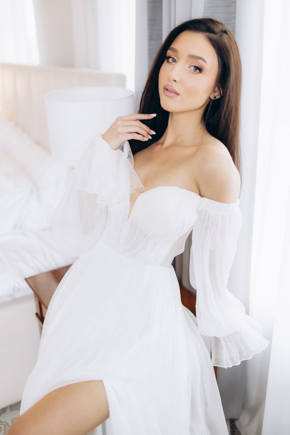 Elegant off-shoulder white dress with flowing sleeves and ruched bodice in a bright, minimalist setting.