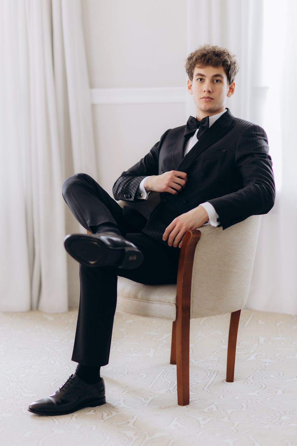 Young man in classic black tuxedo and bow tie poses elegantly in chair against white curtain background.