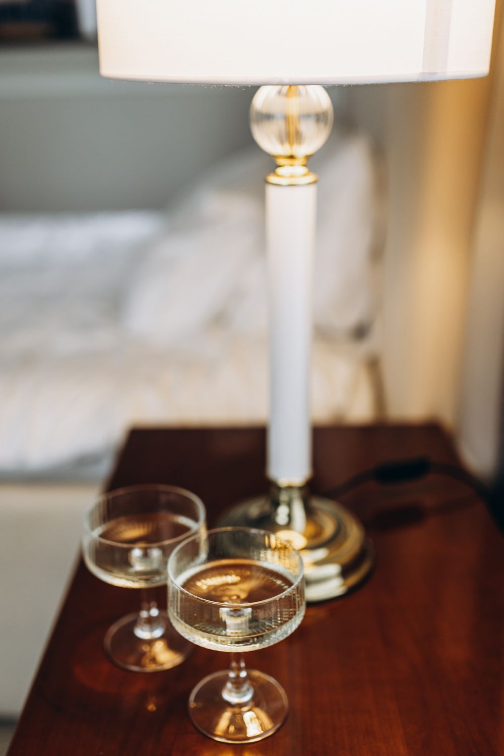 Two elegant champagne glasses on a wooden table next to a white table lamp with brass accents.