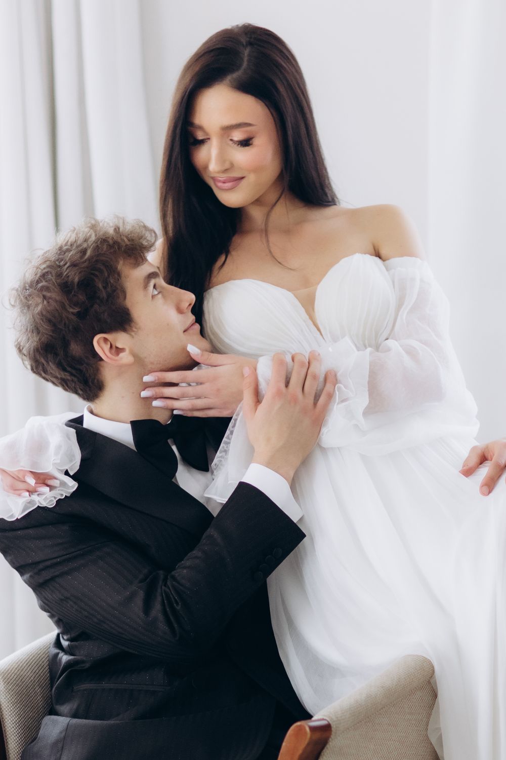 Romantic couple in wedding attire embrace tenderly, with bride in white off-shoulder dress and groom in black suit.