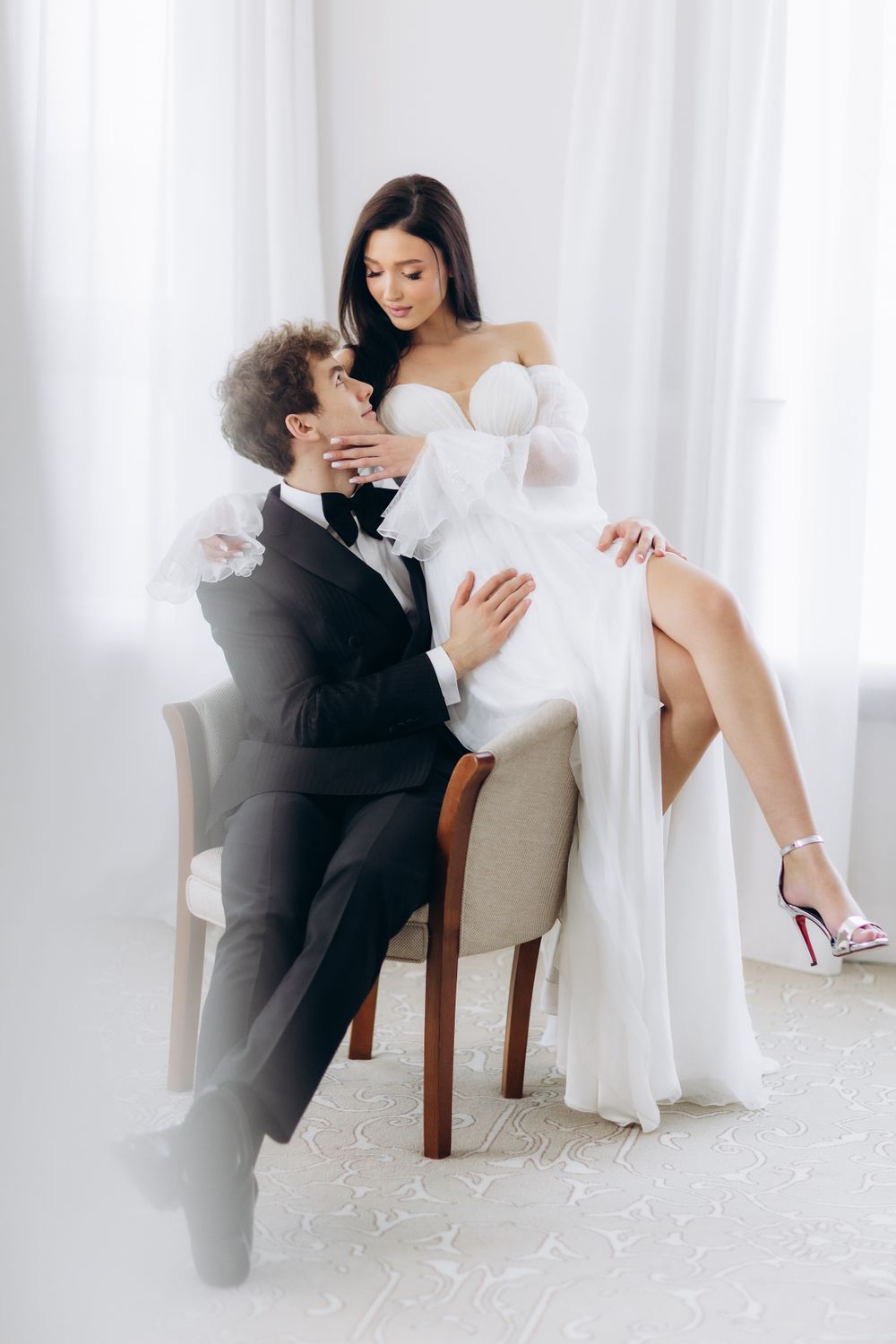 Romantic wedding couple in white and black attire share an intimate moment while seated on beige chair.