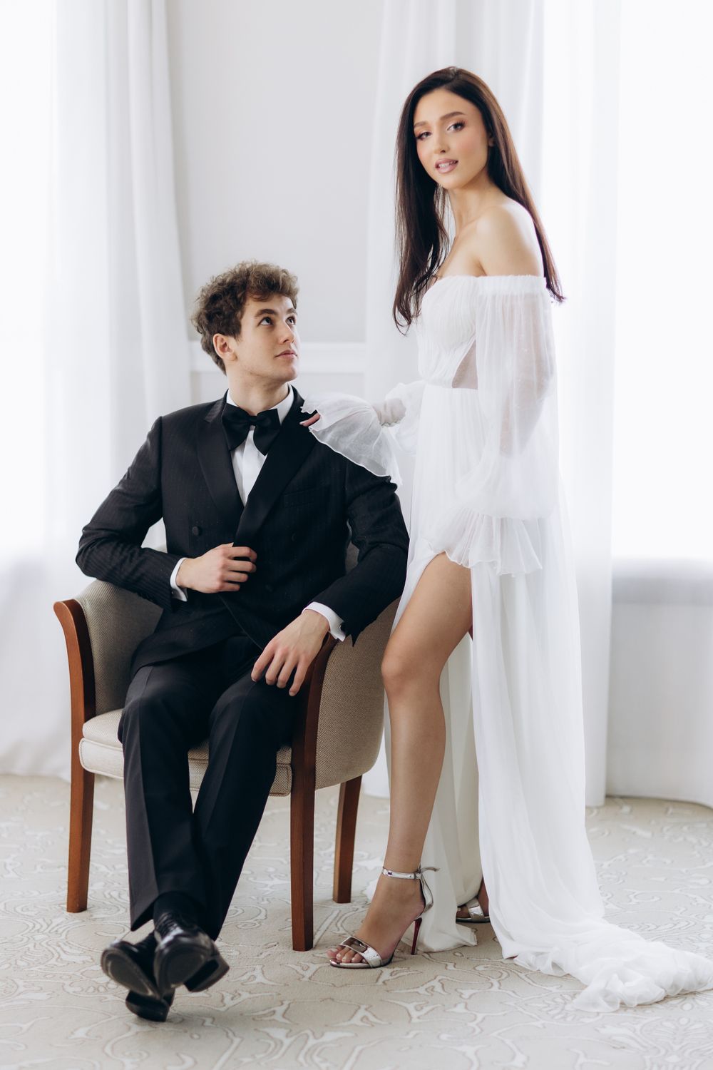 A couple in formal wedding attire poses in a bright white room with the groom seated and bride standing beside him.