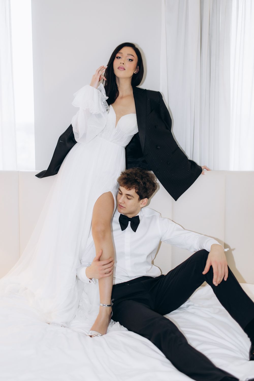 Stylish couple in black and white formal wear pose artistically against white curtain backdrop for wedding photo.