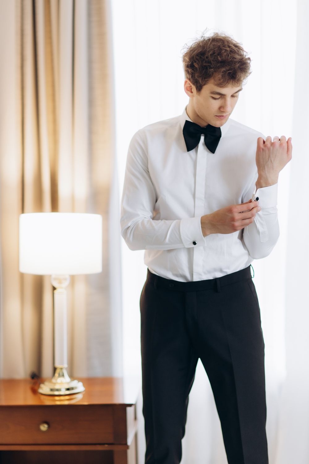Young person in formal attire with black bow tie adjusts white dress shirt sleeve cuff next to table lamp.
