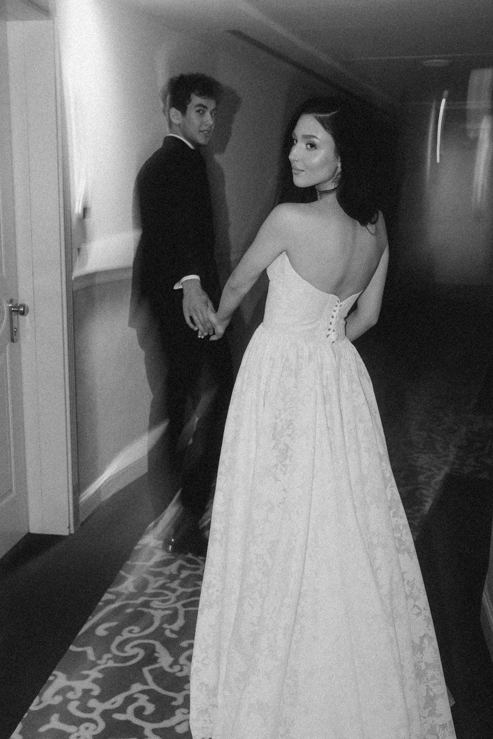 Black and white photo of couple in formal attire walking down a hallway, showing back of elegant white gown.