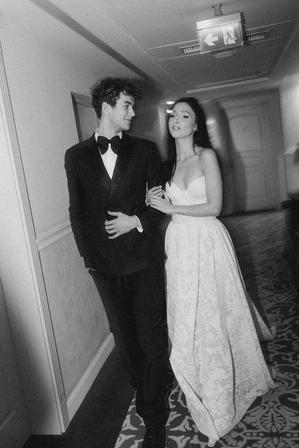 A couple in formal attire stands together in an elegant hallway, with the woman wearing a white lace gown.