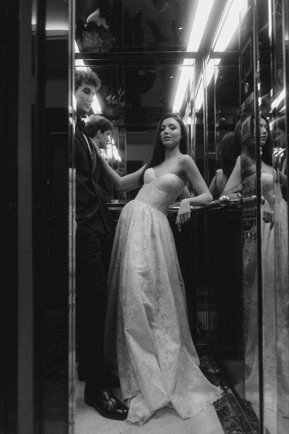 A black and white photo of a figure in an elegant white ball gown standing in a narrow hallway under fluorescent lights.