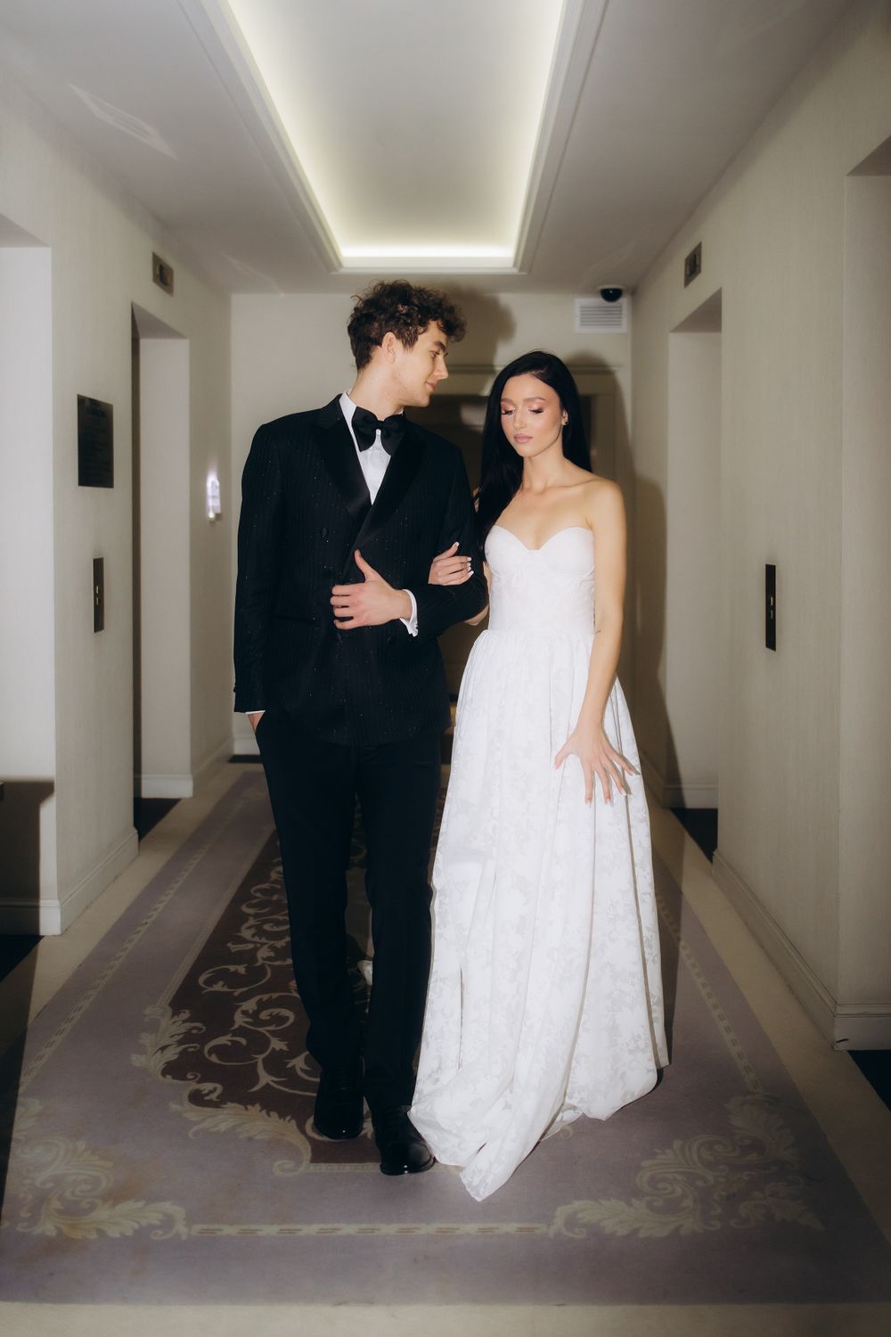 A bride in a white gown and groom in black tuxedo stand together in an elegant hallway with soft lighting.