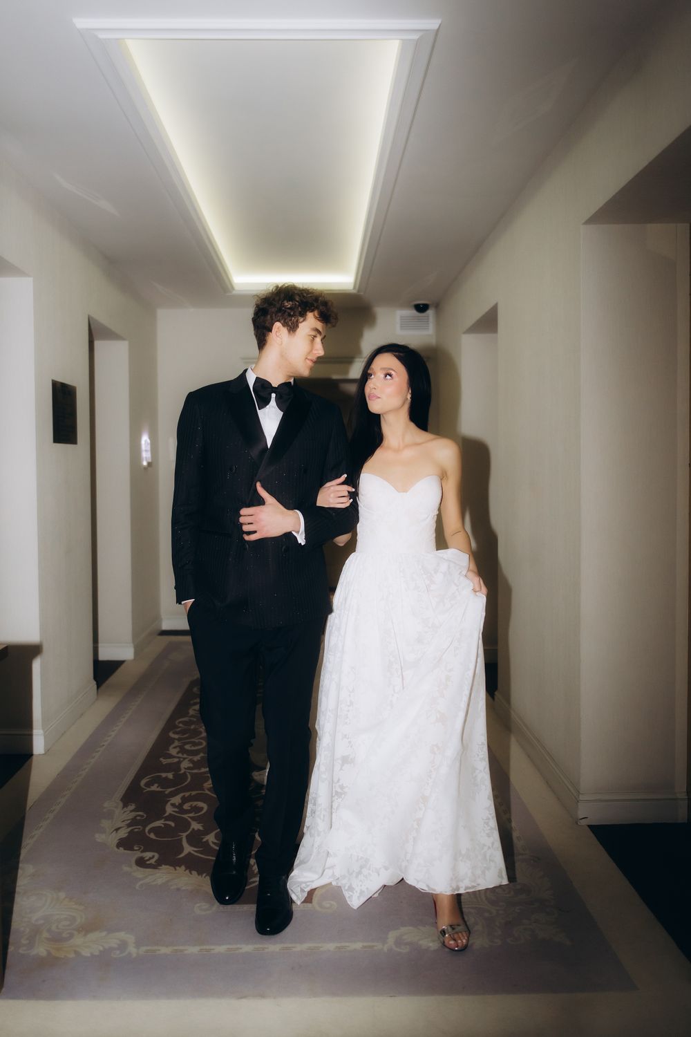 An elegant couple in formal wedding attire walks together in a hotel hallway with modern lighting.