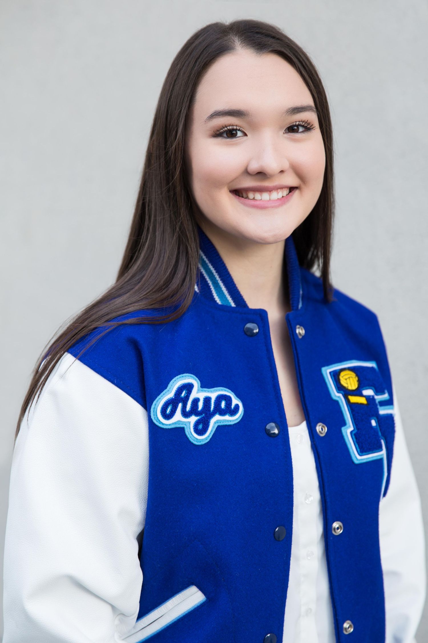 Student wearing a blue and white varsity letterman jacket smiles warmly against a light gray background.