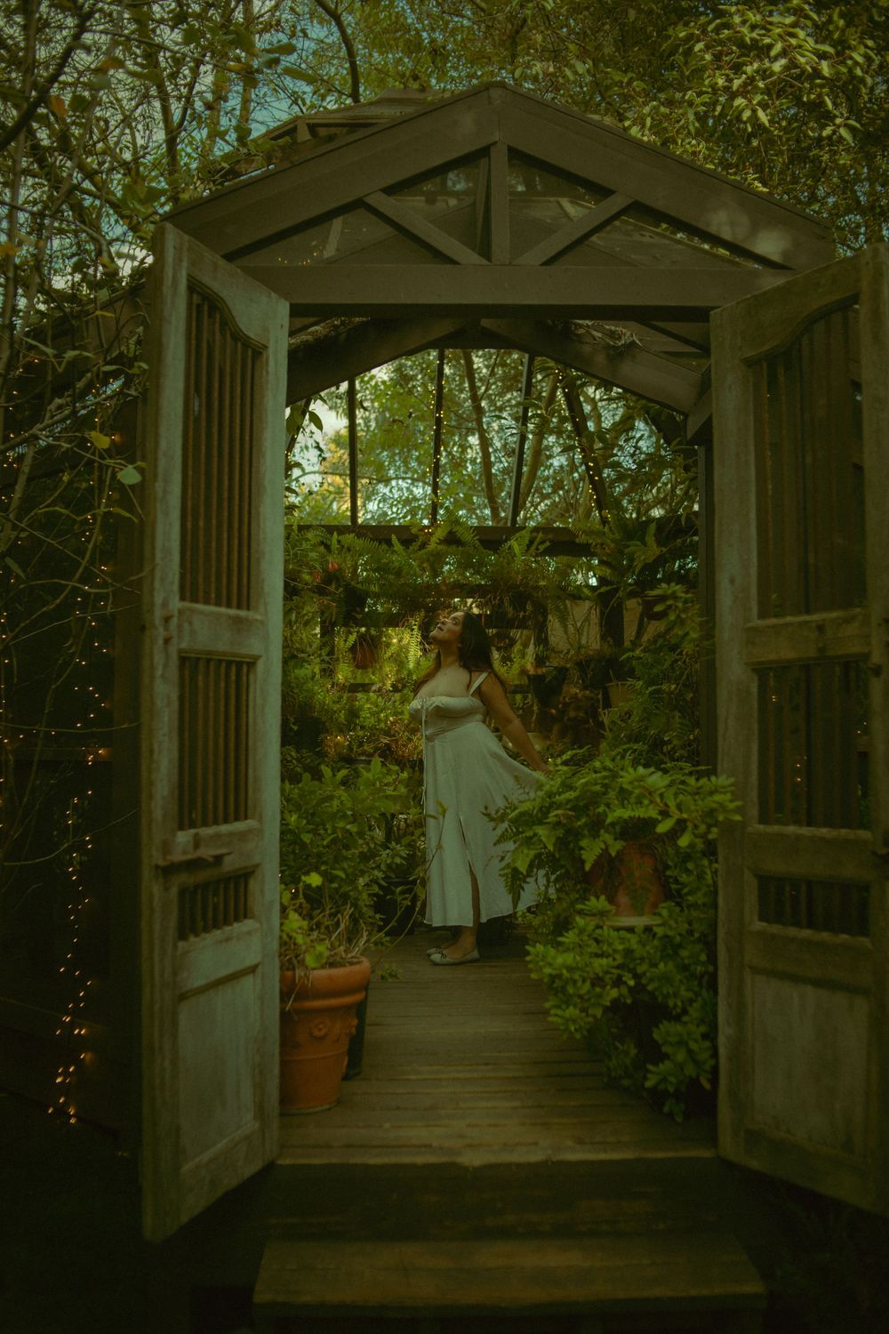 Photography of wooden garden gate entrance frames a path through lush greenhouse with a woman in white dancing