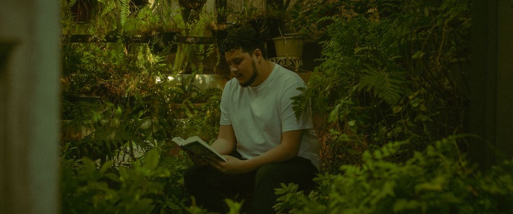 Photograph of a man sits reading in a lush greenhouse in San Marcos California