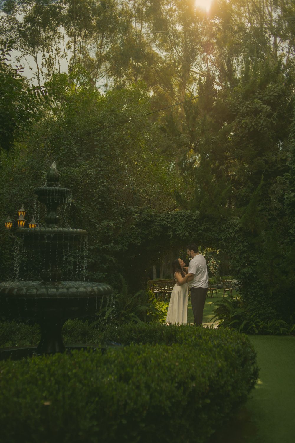 A romantic garden photography with a tiered fountain and engaged couple embraced in golden sunlight filtering through trees at dusk.