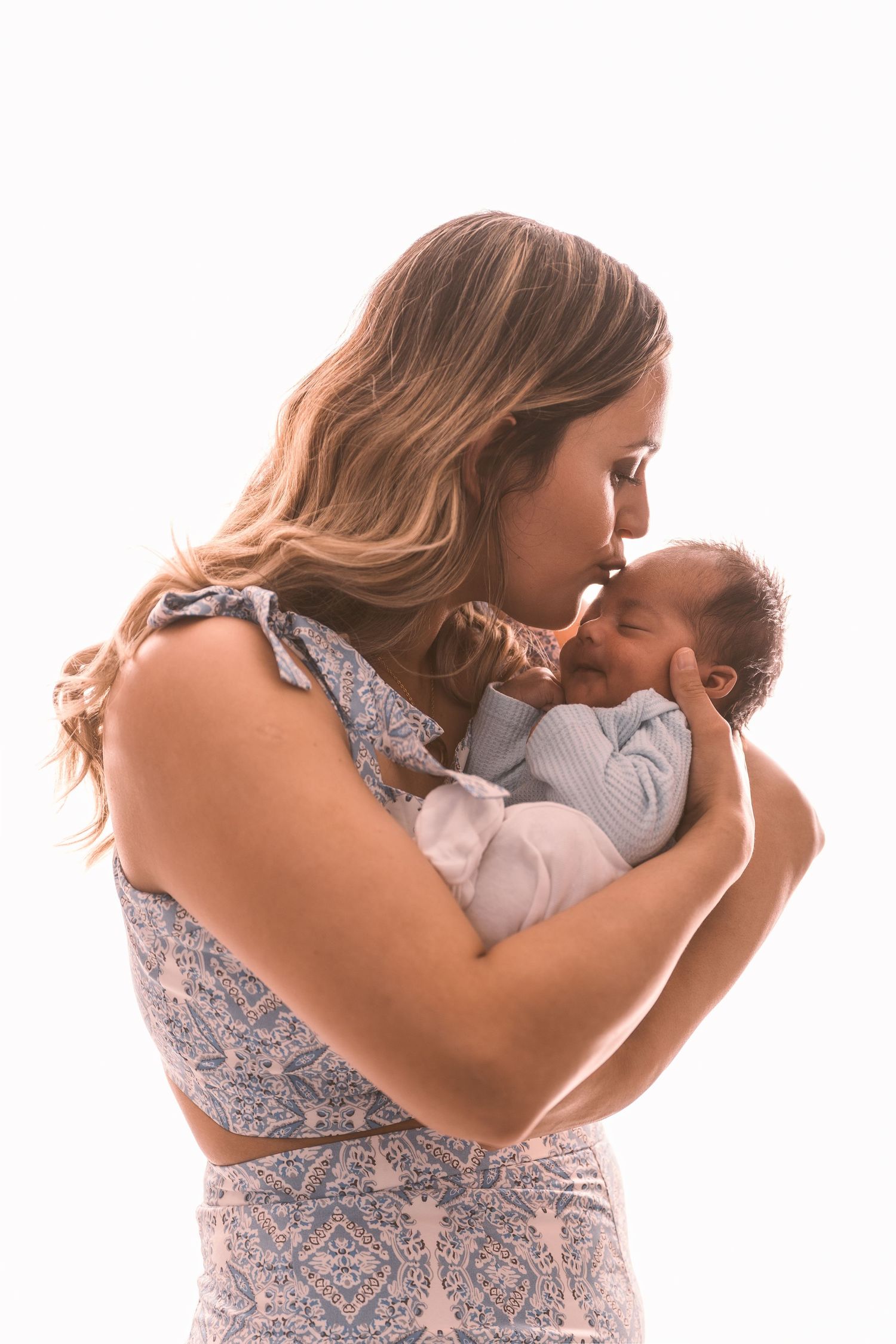 Woman in floral dress holding and kissing baby against white background.