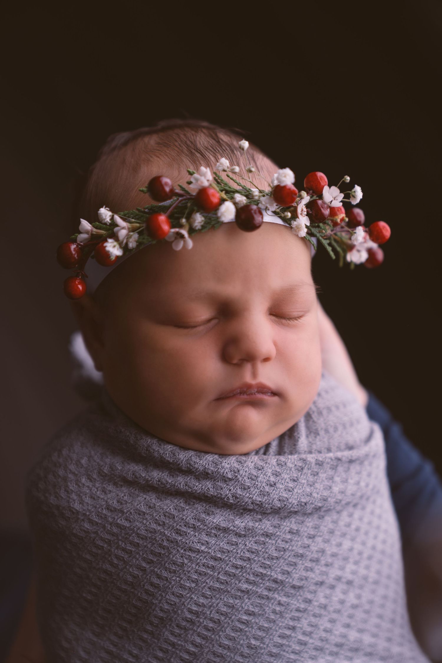 Sleeping newborn wrapped in gray swaddle wearing delicate floral crown with red berries against dark background.