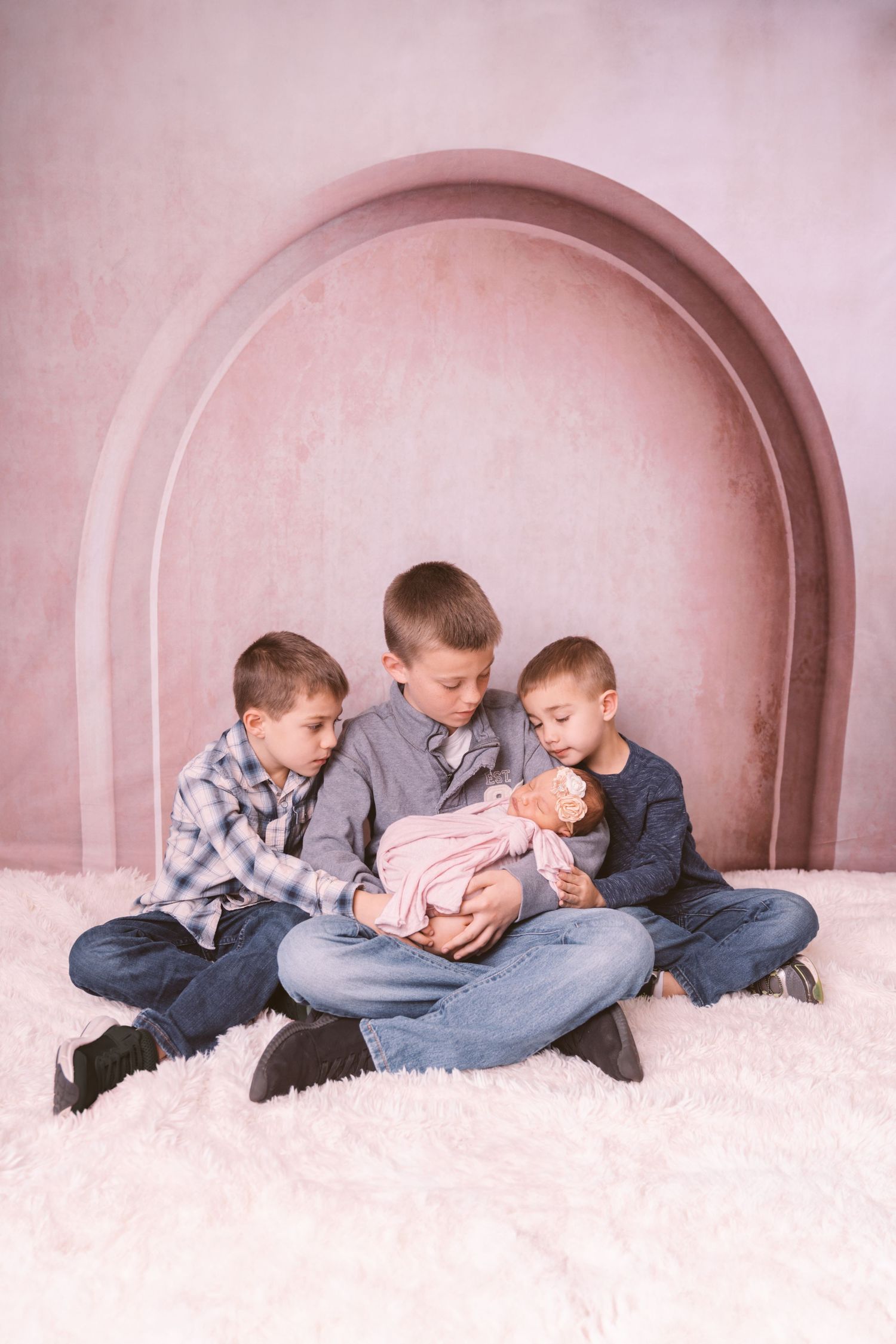 Three siblings lying together on plush carpet looking at newborn in pink alcove setting.