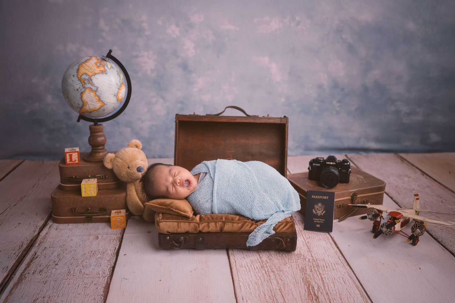 Vintage-styled newborn photo with globe, suitcase and travel props on wooden floor.