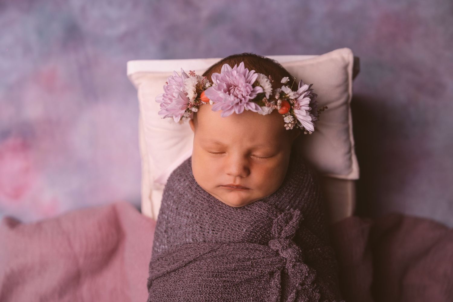 Sleeping newborn in gray wrap with pink floral headpiece against purple backdrop.