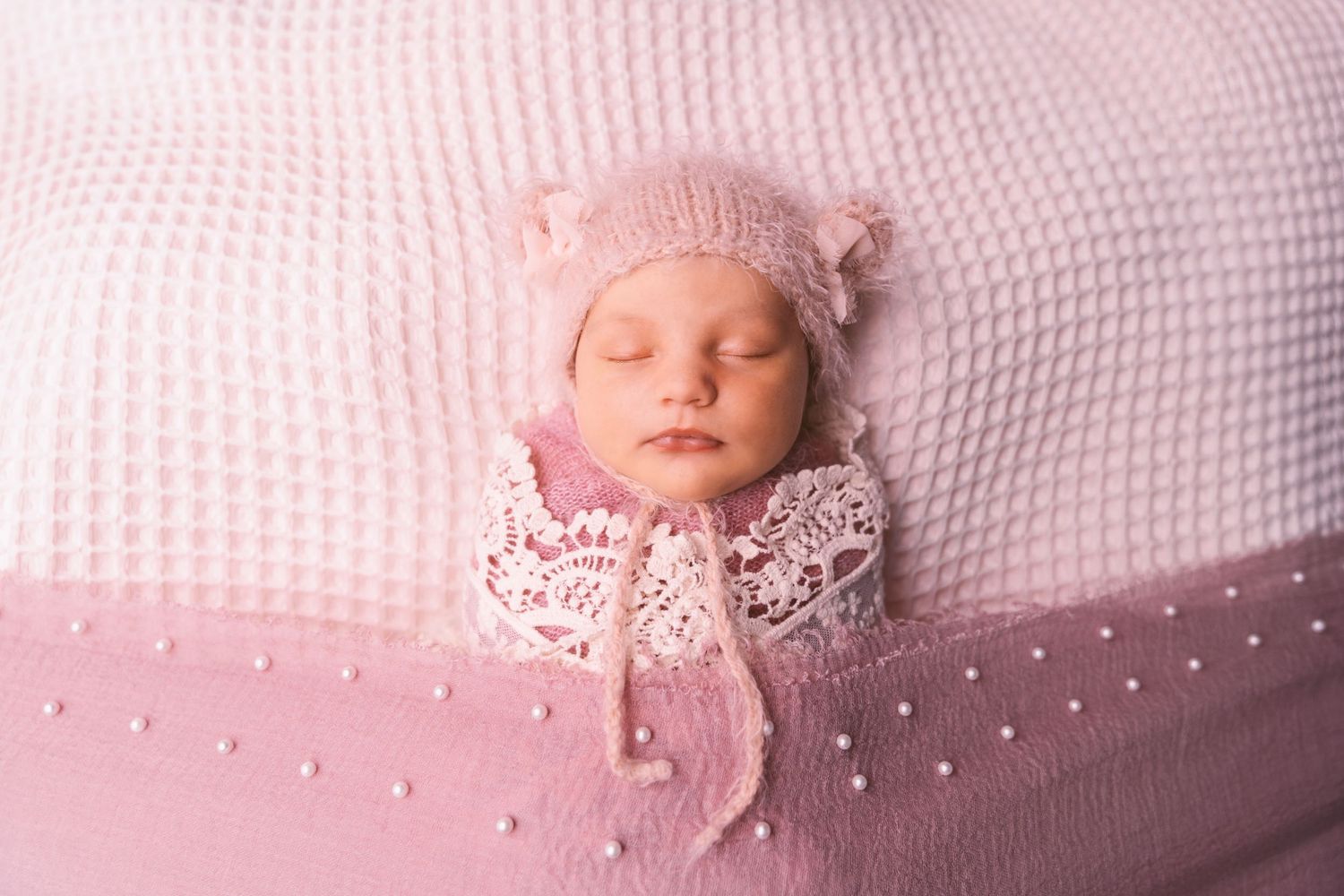 Newborn sleeping on textured white blanket with pink bow and lace accessories.