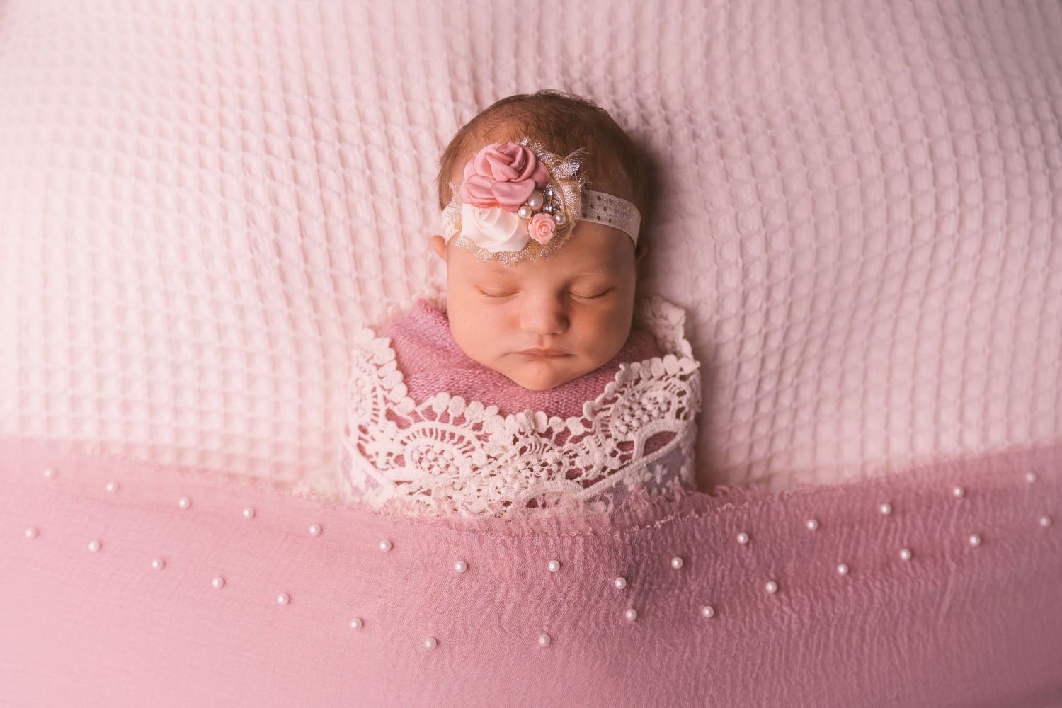 Sleeping newborn on white textured blanket with pink lace accessories.
