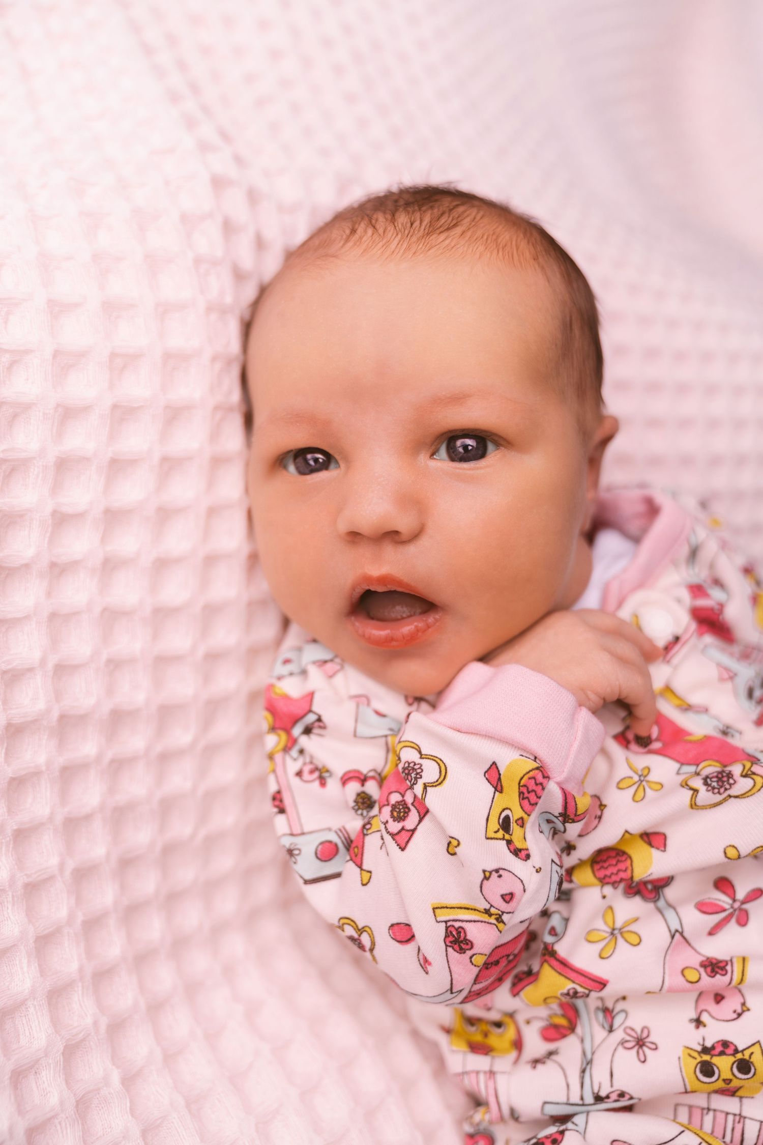 Baby wearing pink patterned clothing lying on textured pink blanket.