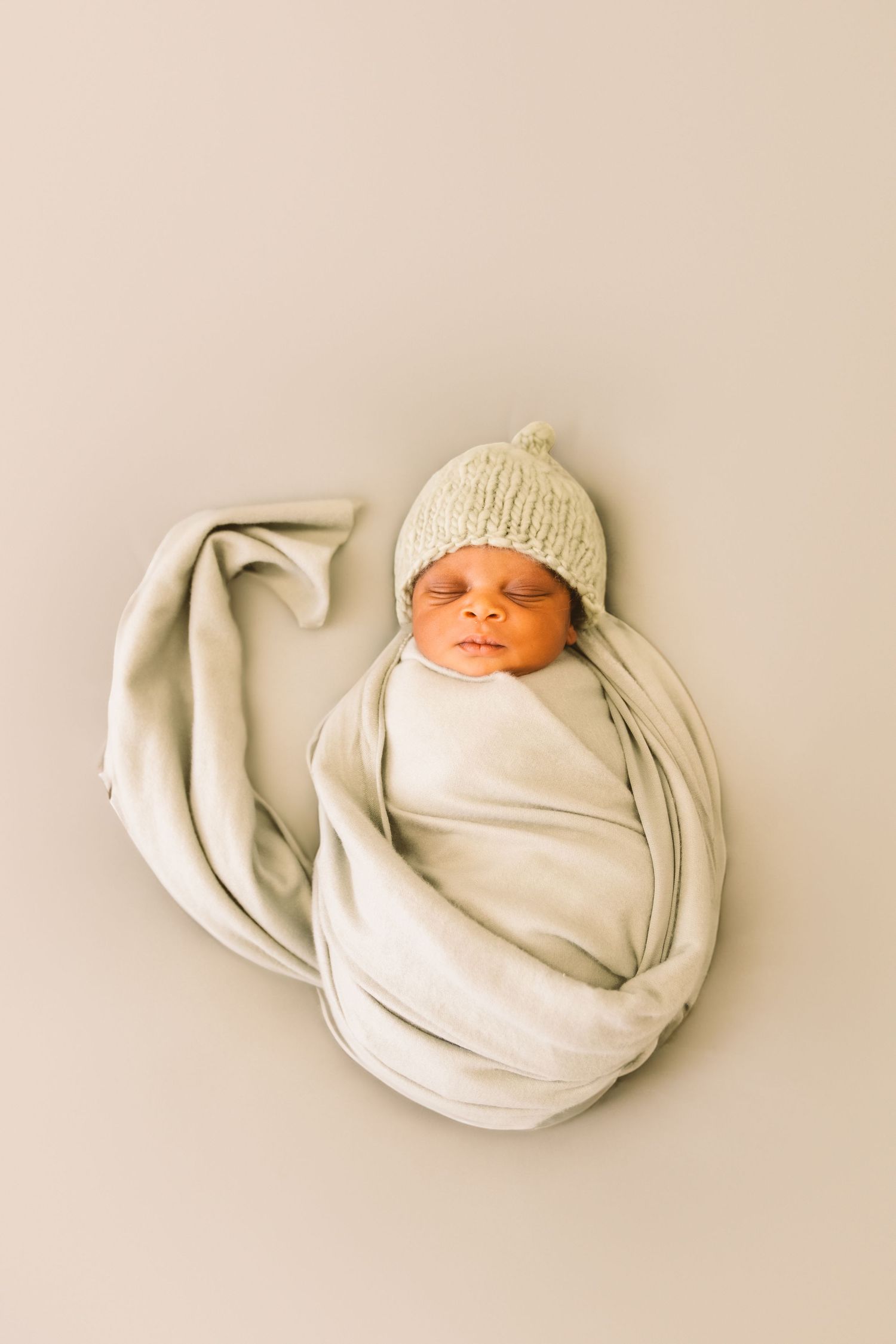 Newborn baby wrapped in cream swaddle wearing knit hat on beige background.