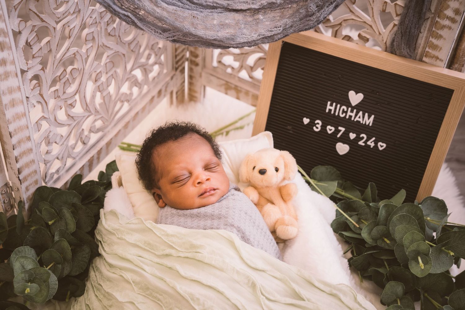 Sleeping newborn in gray swaddle with teddy bear and decorative greenery.