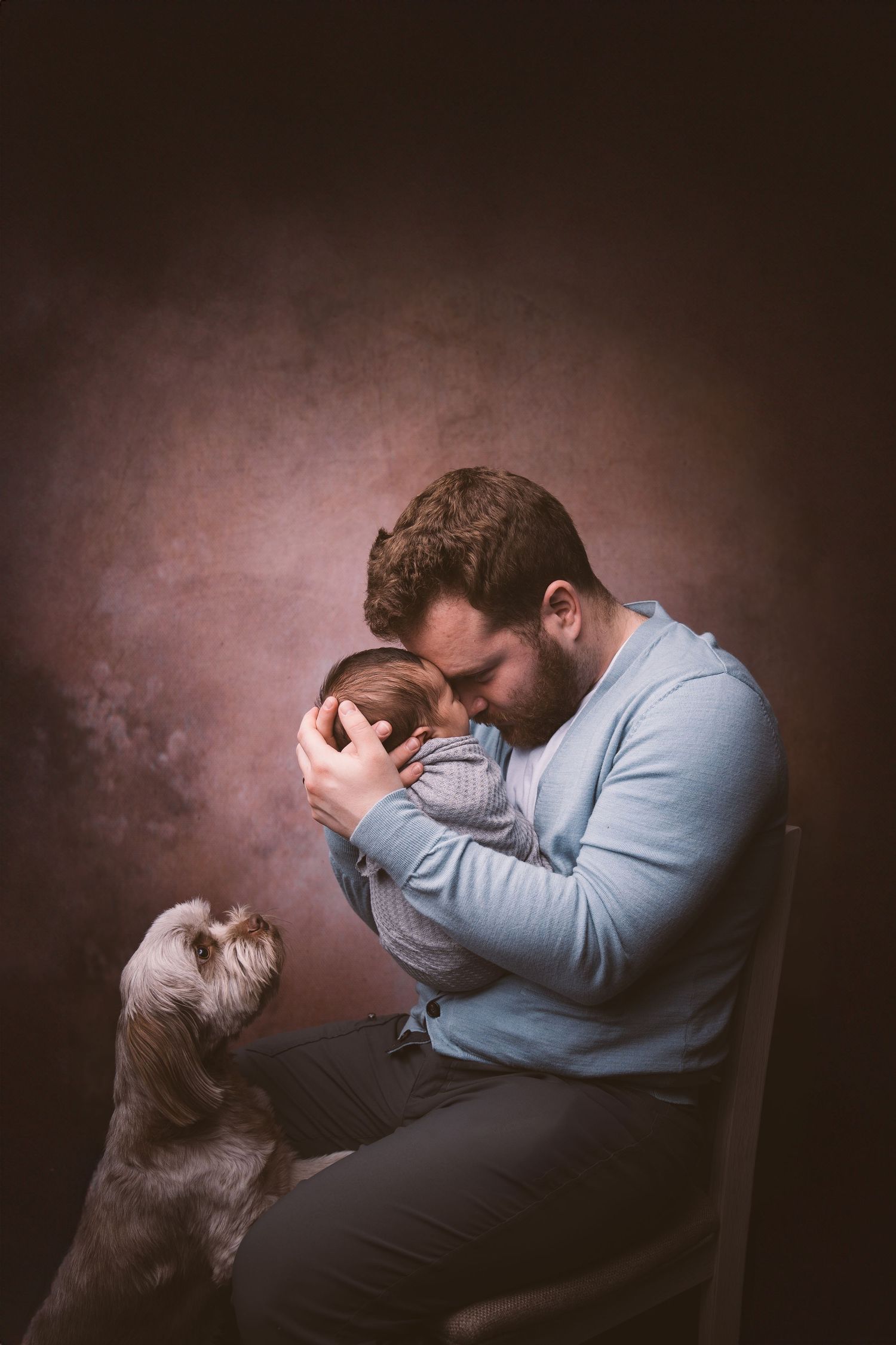 Person in blue sweater sharing intimate moment with baby while cat looks on curiously.