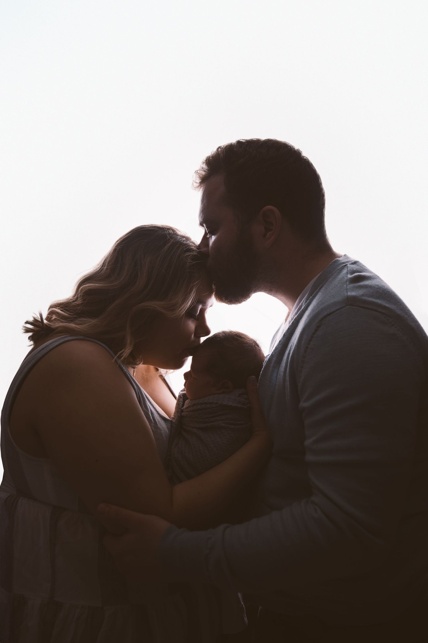 Silhouetted family members sharing intimate moment with newborn against bright background.