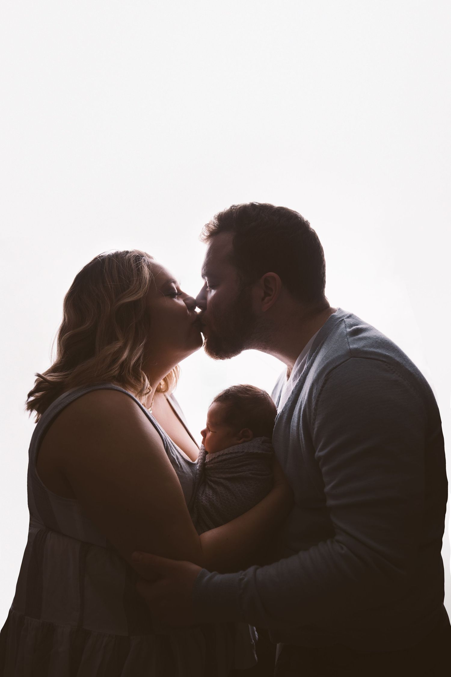 Family members in silhouette embracing and bonding with newborn against white backdrop.