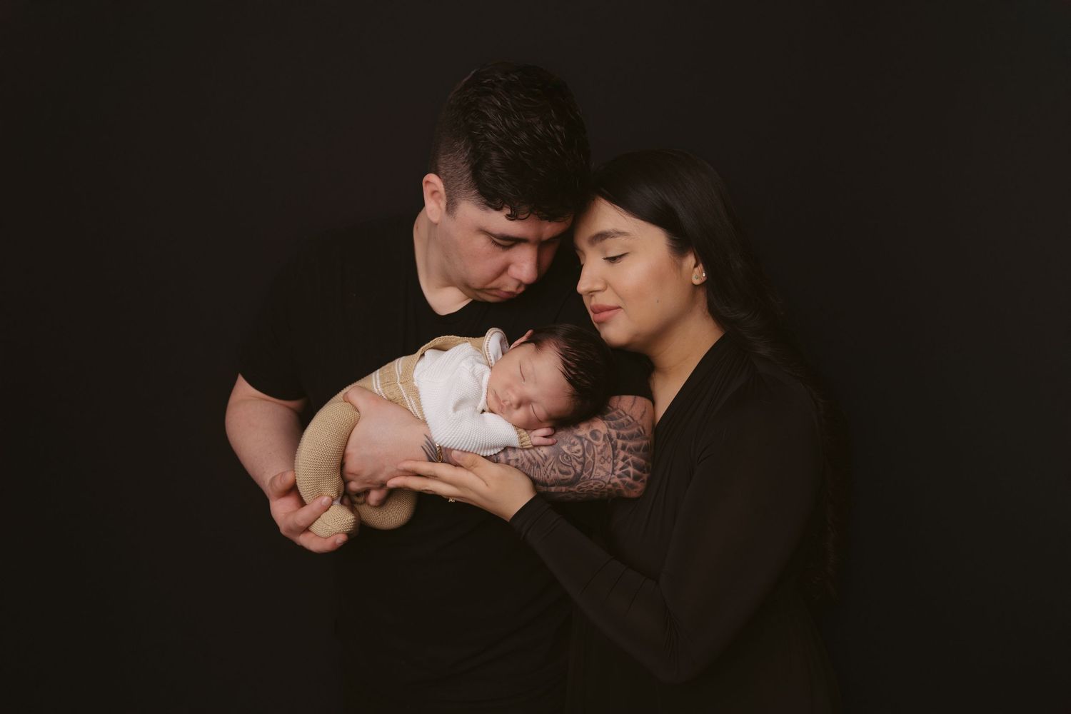 Family portrait against black background with sleeping baby in white clothing.