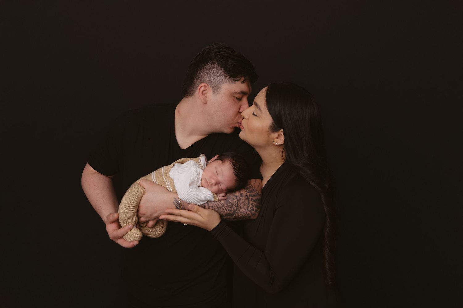 Tender family moment captured against dark background with sleeping infant.