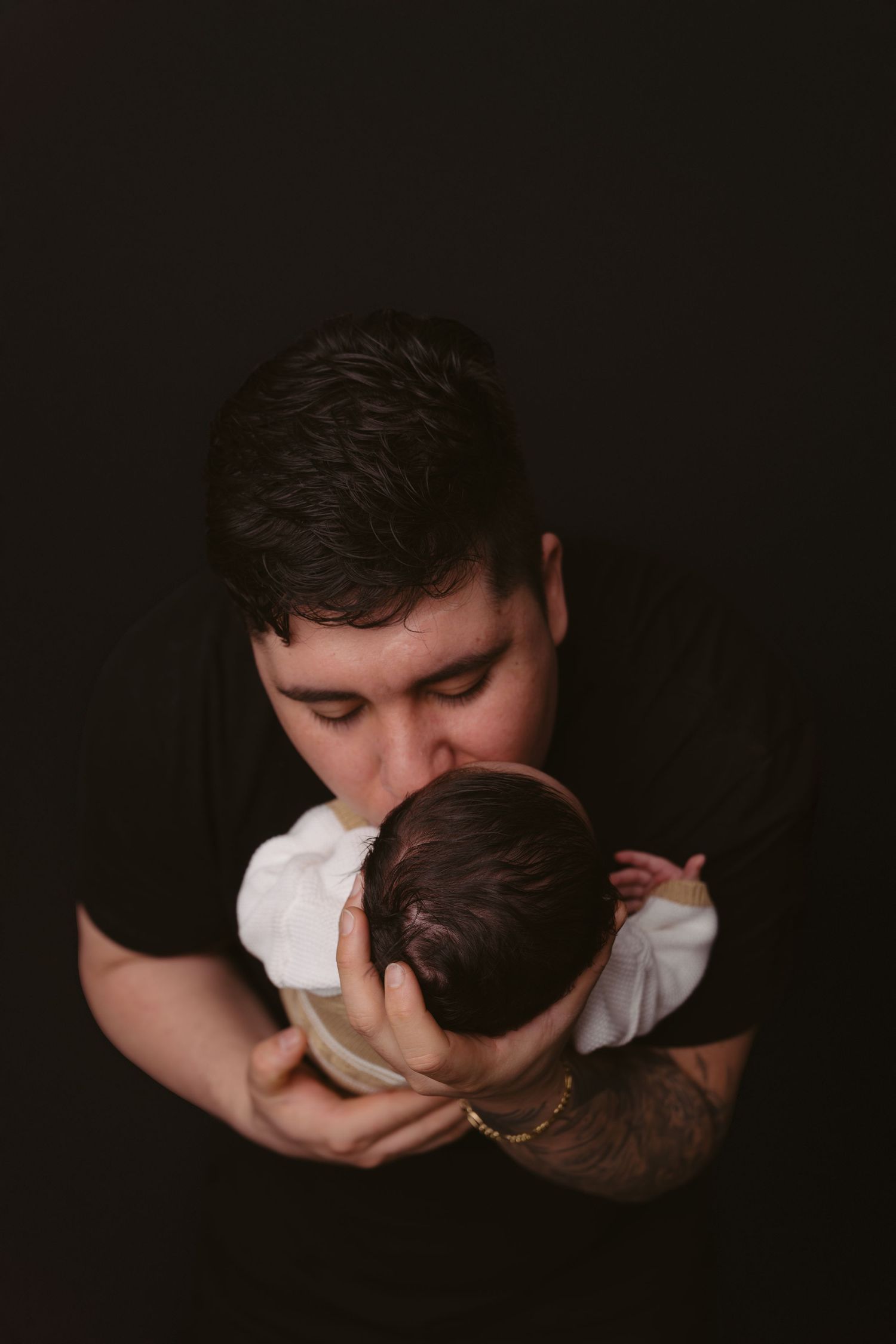 Parent holding sleeping infant in white onesie against dark backdrop.