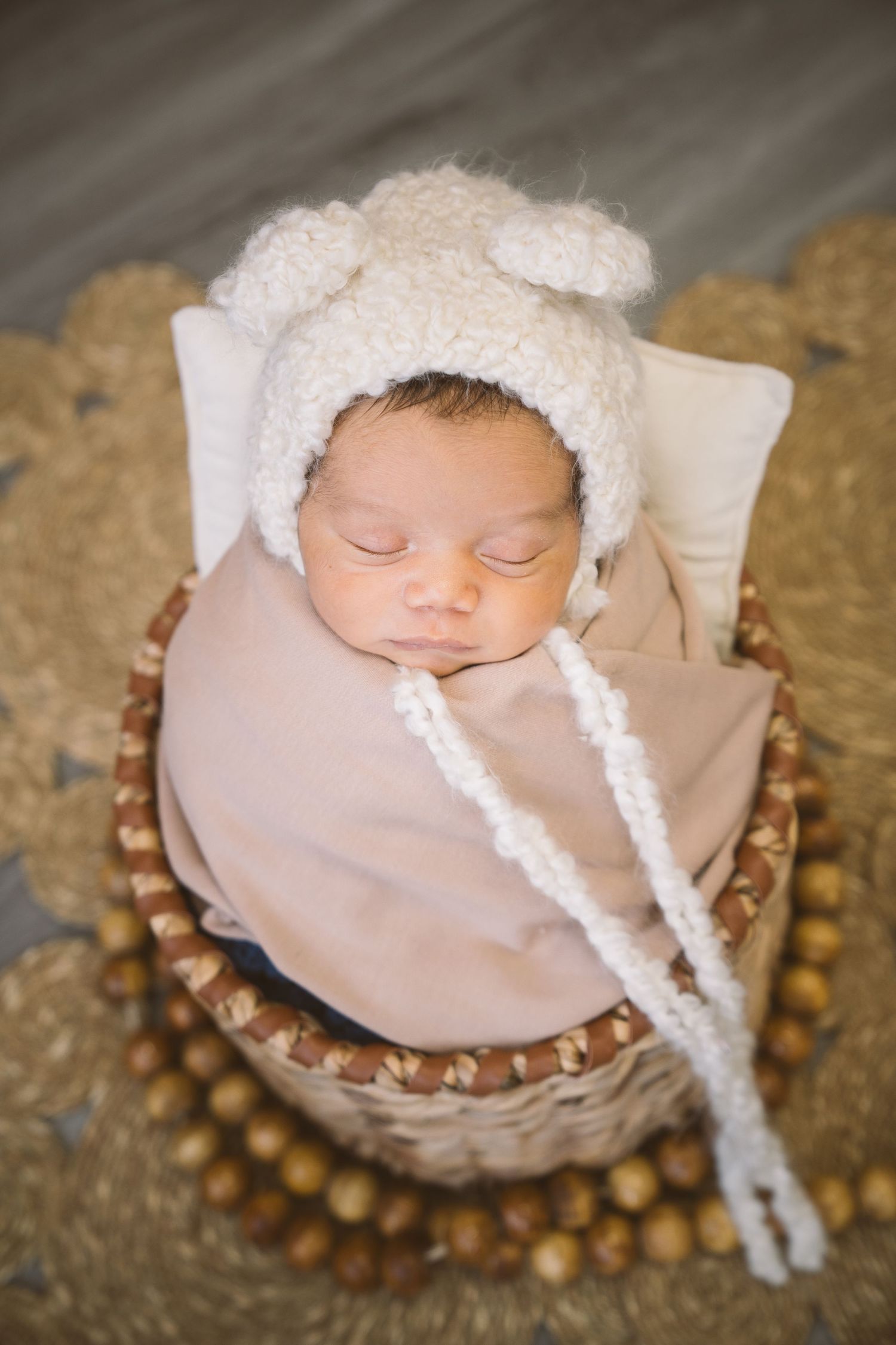 Sleeping newborn baby wearing a white knit hat in a rustic basket with beaded decorations.