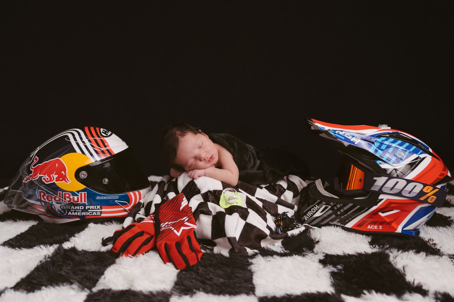 Racing themed newborn photo with motorsport helmets and checkered flag background.