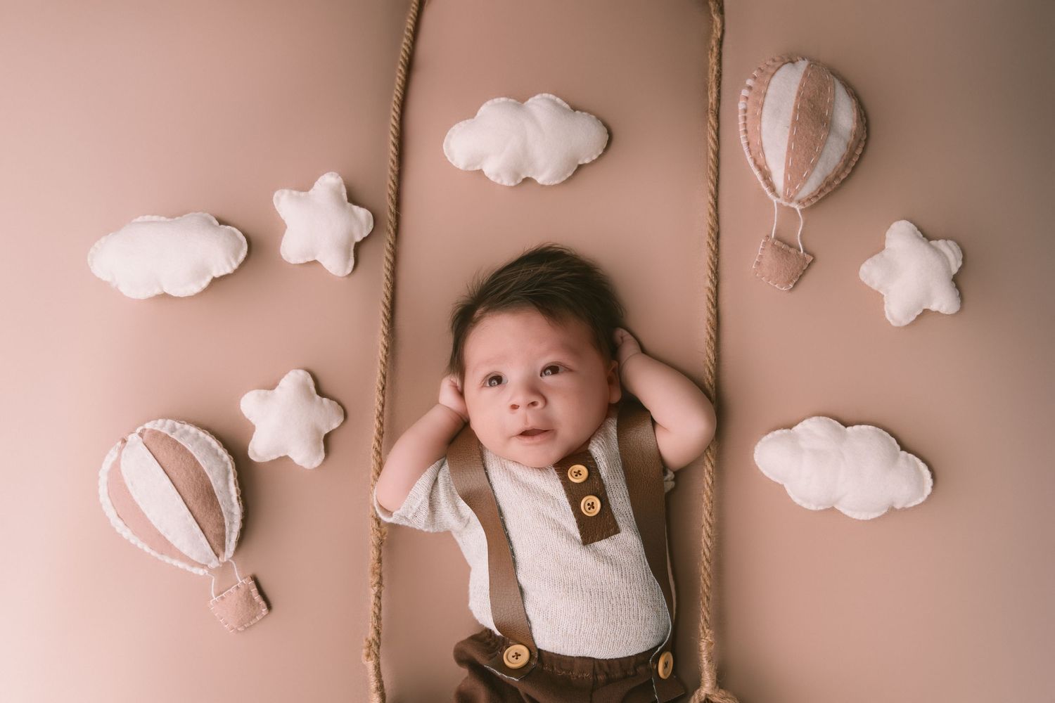 A baby lies on pink backdrop with decorative white clouds and hot air balloons.