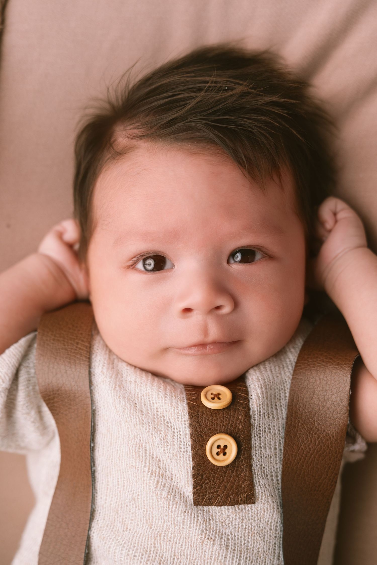 A baby wearing a tan suspender outfit smiles while lying on pink fabric.
