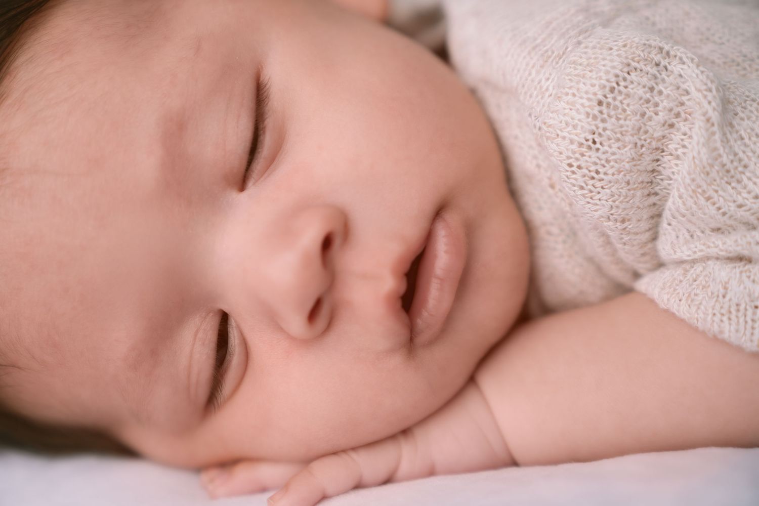 Peaceful sleeping newborn wrapped in cozy white knit blanket on pink backdrop.