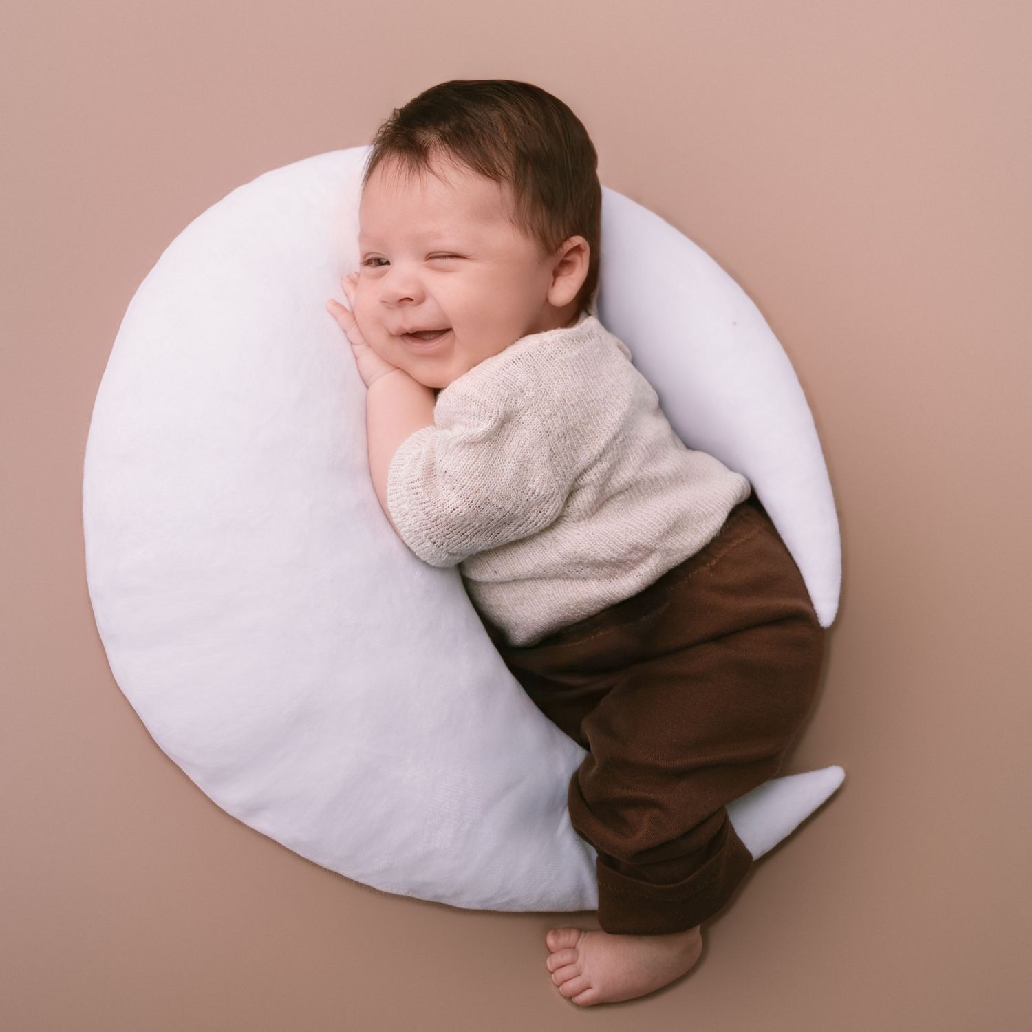 Sweet baby in brown and white clothing smiling while lying on white pillow.