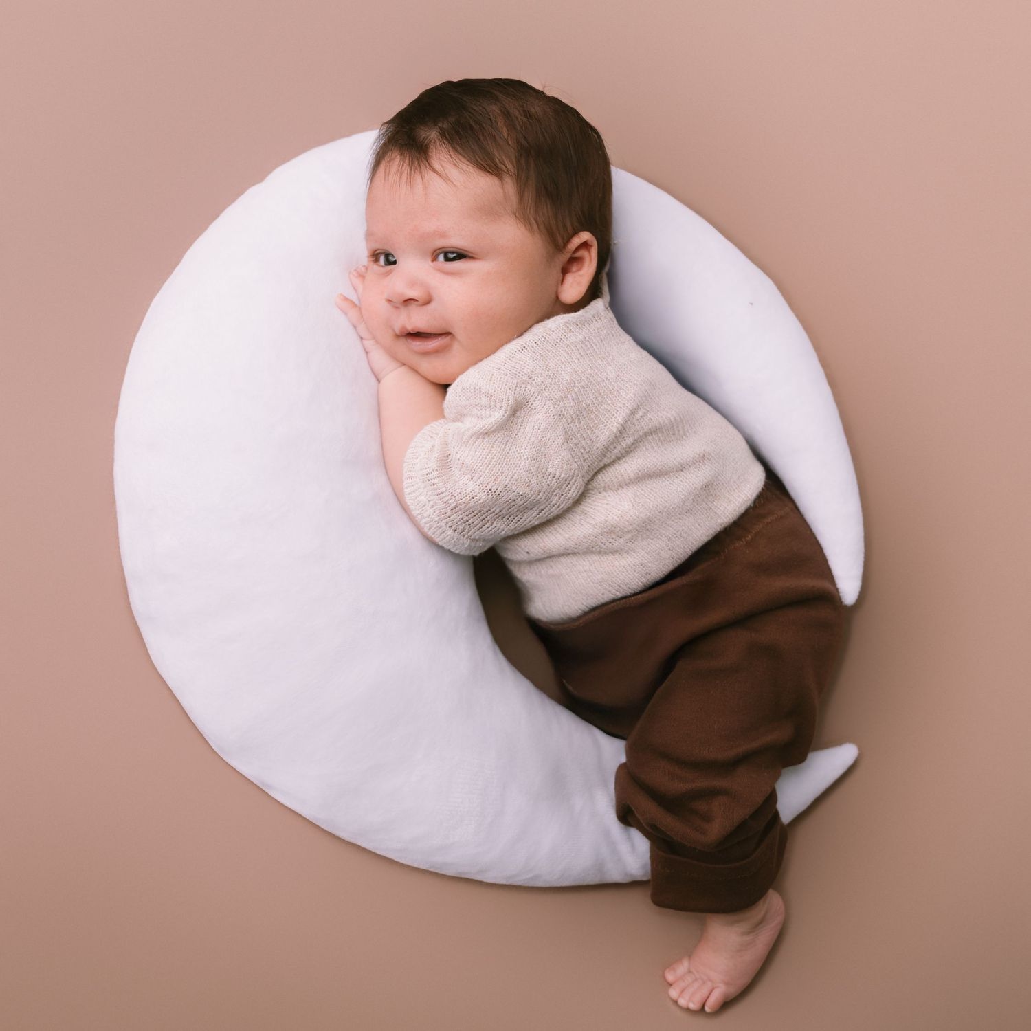 Newborn baby wearing brown and white outfit resting on white cushion against pink background.