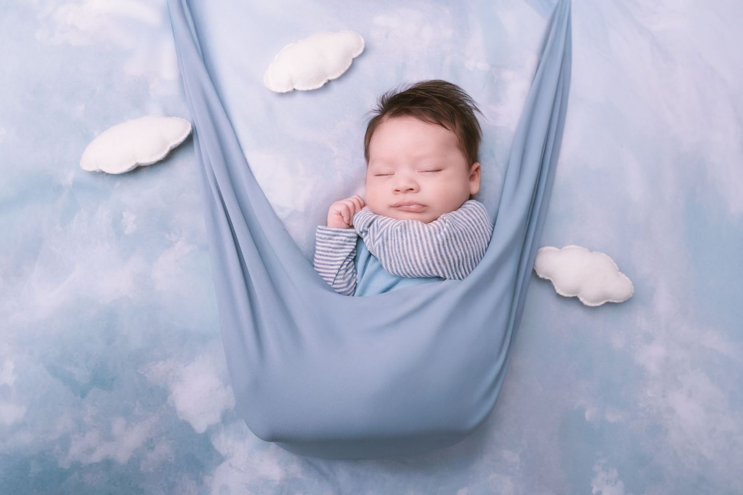 Dreamy newborn portrait in blue fabric hammock with white cloud decorations.