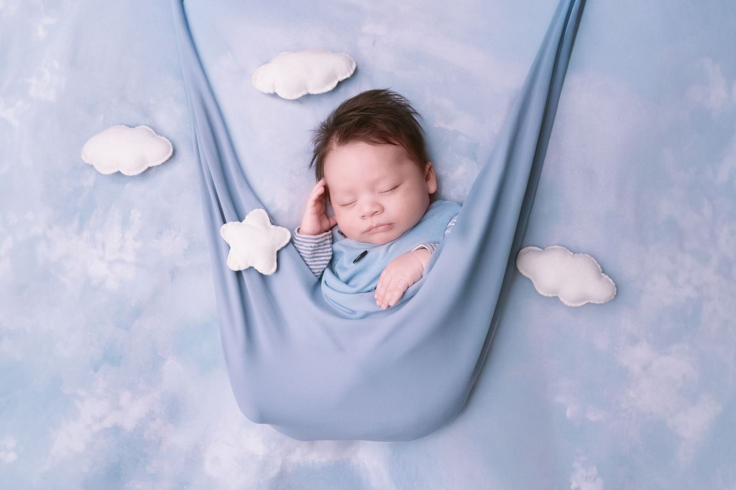 Baby sleeping peacefully in blue fabric sling with white cloud accents.