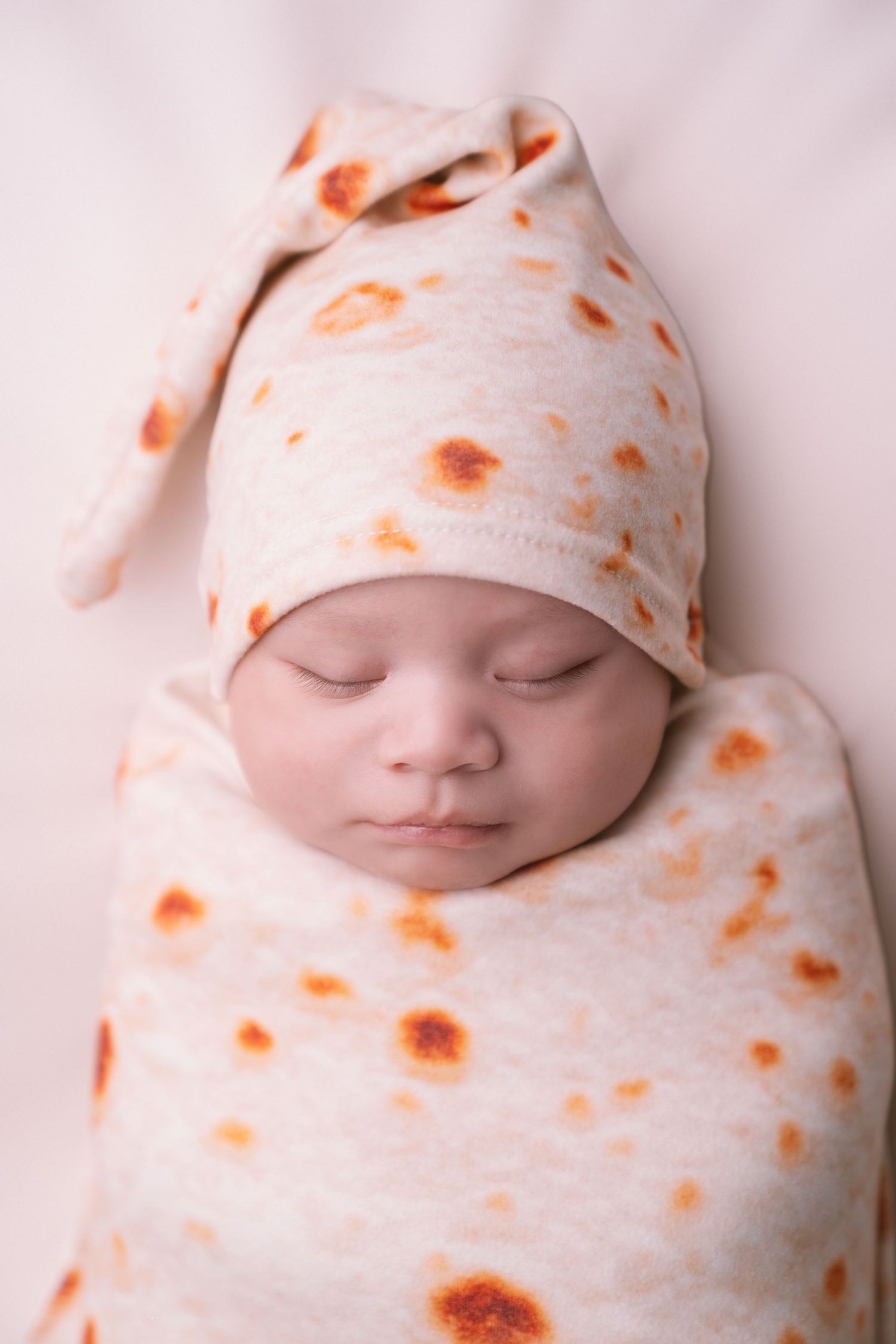 Sleeping newborn wrapped in tortilla-patterned swaddle blanket on white backdrop.