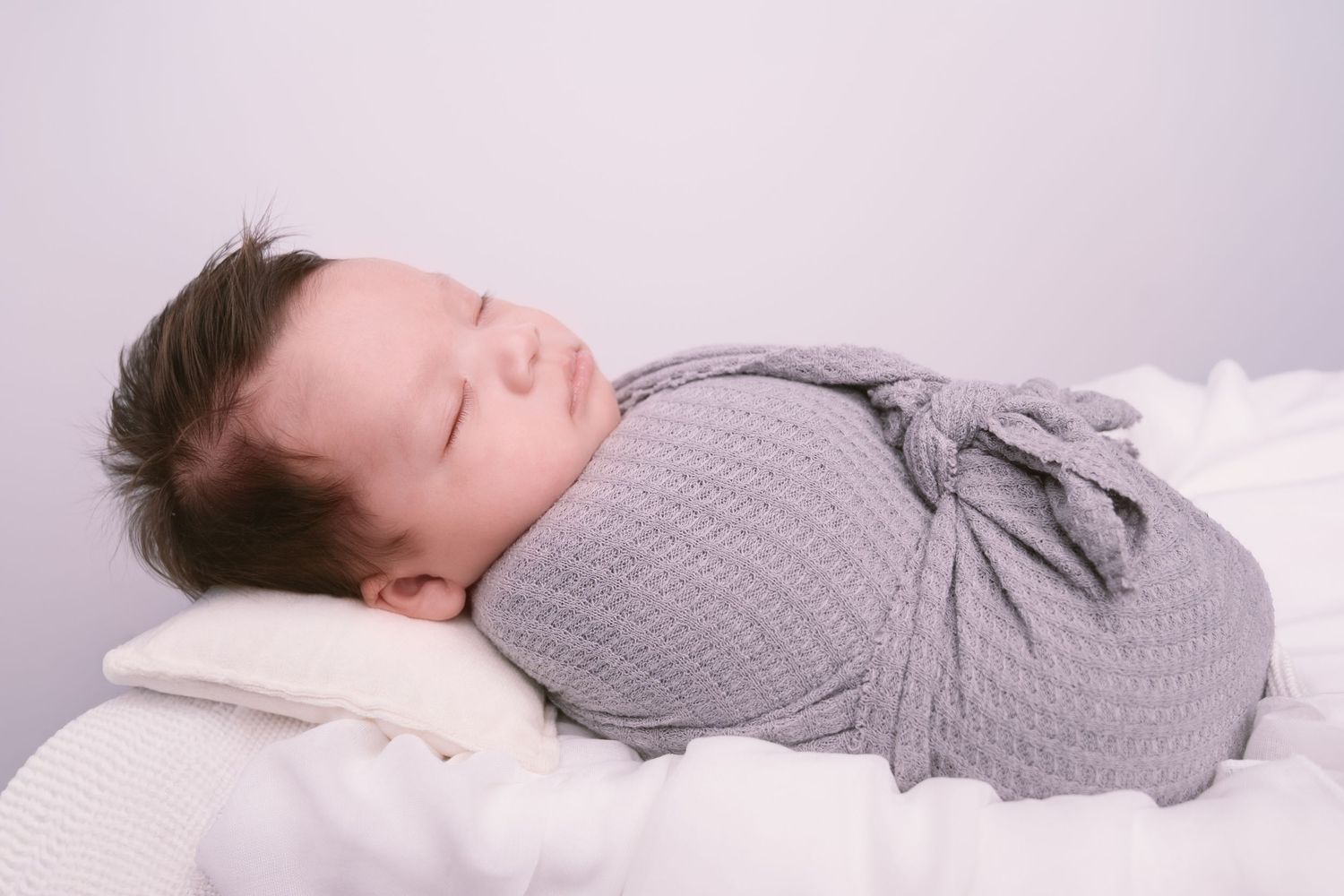 Side view of sleeping newborn wrapped in gray knit blanket on white surface.