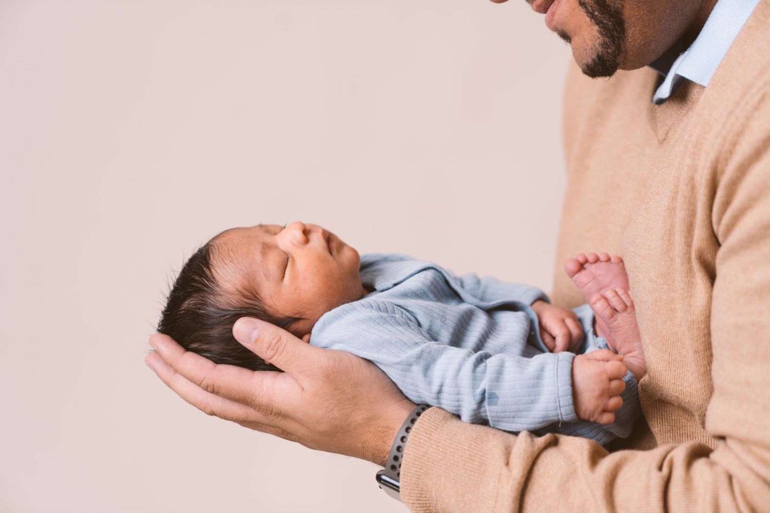 Newborn baby in light blue outfit being held carefully against beige background.