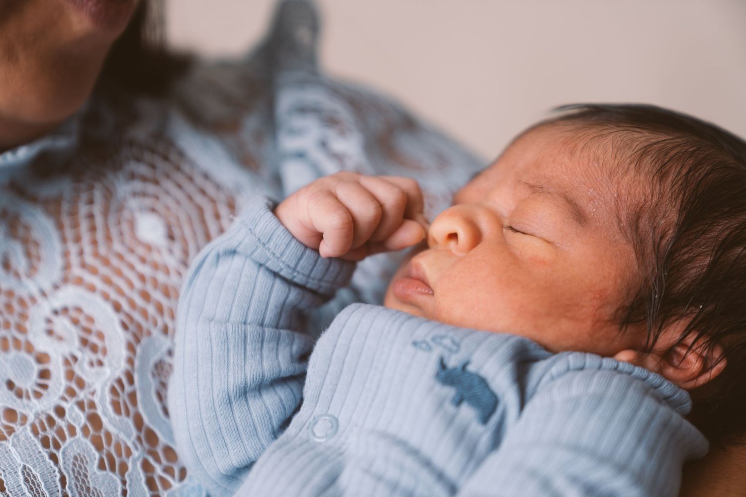 Gentle hands cradle a sleeping newborn baby wearing light blue clothing against lace background.