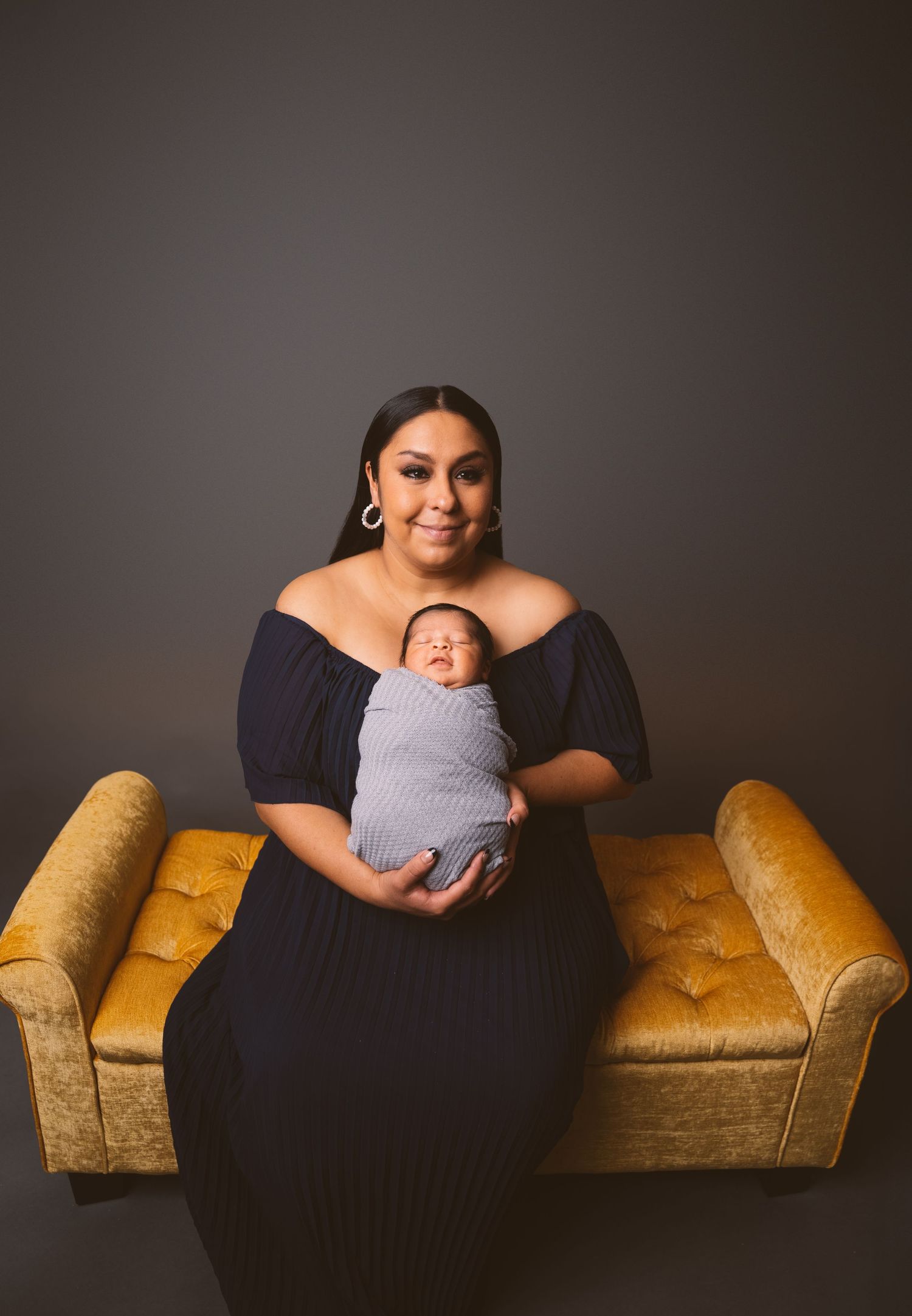 Professional studio portrait on yellow vintage couch with sleeping newborn in gray wrap.